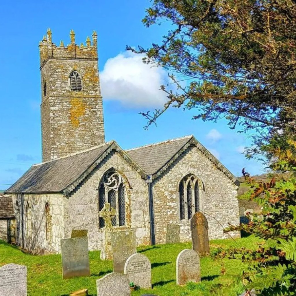 St Adwena’s church and tower at Advent, seen from the green churchyard with gravestones under a bright blue sky