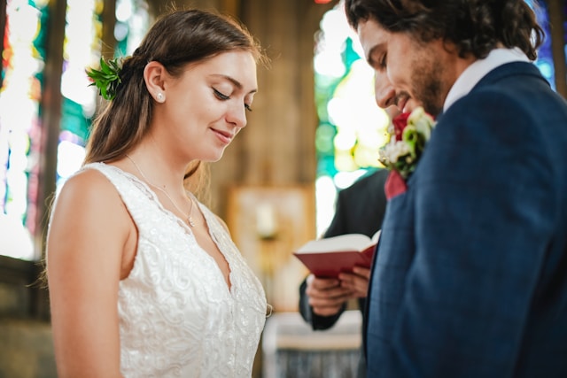 A bride and groom at the front of a church during their wedding or blessing, with stained glass behind them